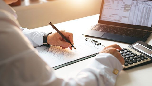 Un hombre está escribiendo en un portapapeles mientras usa una computadora portátil y una calculadora.