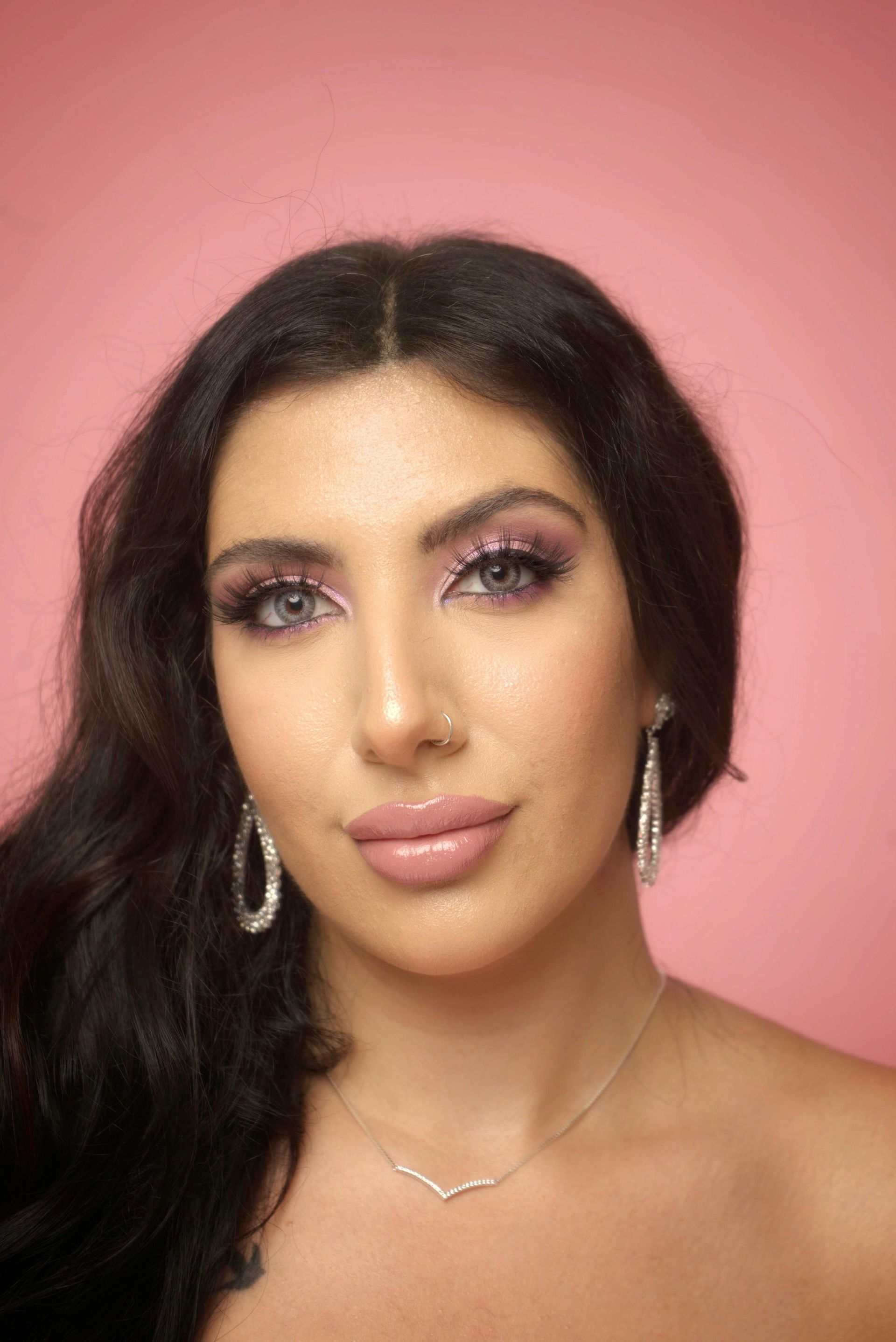 A woman wearing a necklace and earrings is standing in front of a pink background.