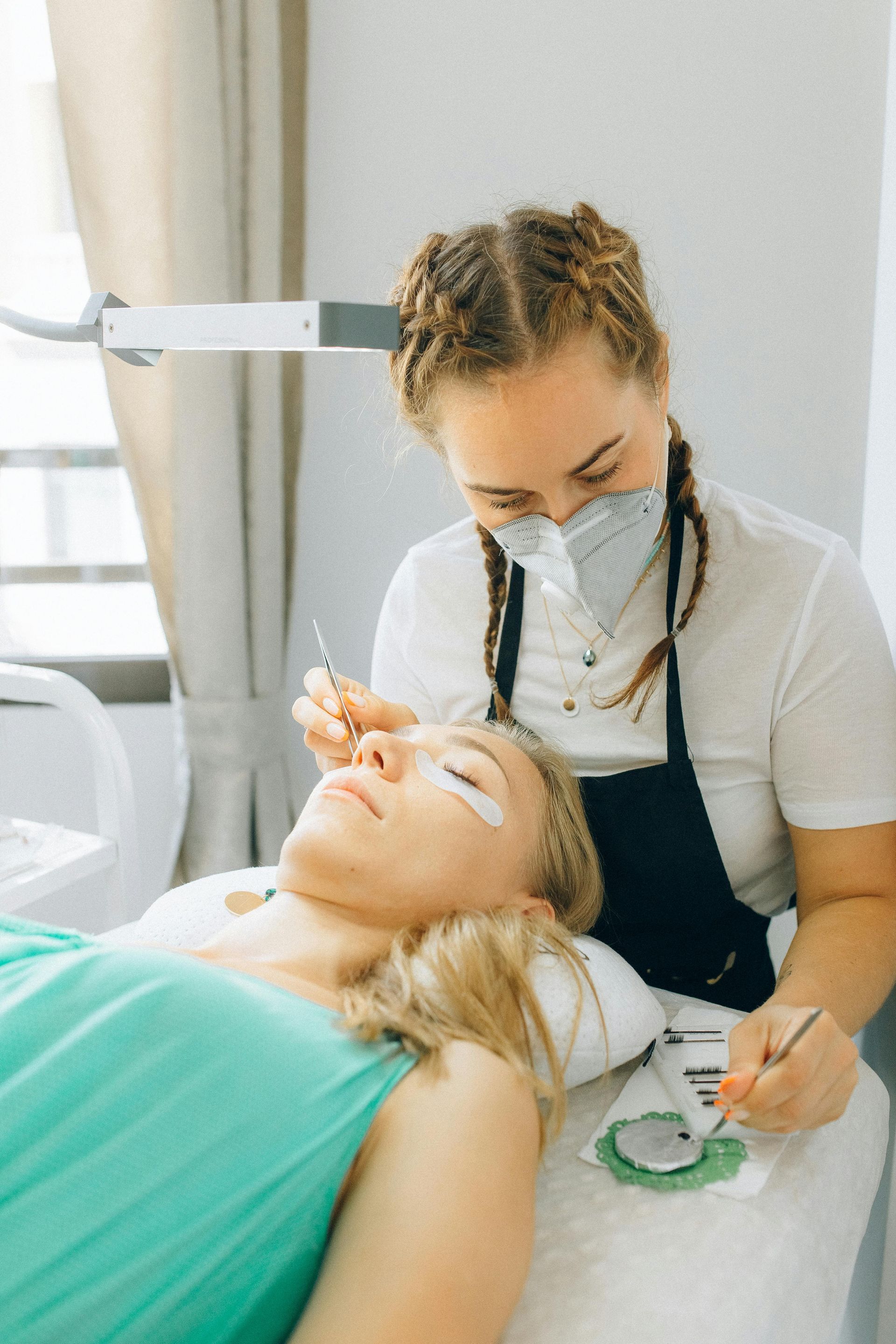 A woman is getting her eyelashes done by a woman wearing a mask.