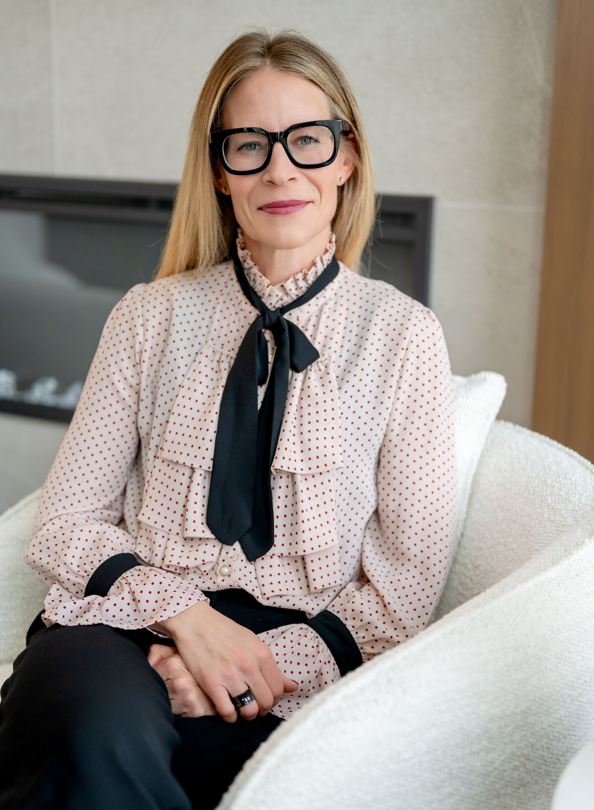 Woman in glasses and blouse with tie, sitting in a white chair and smiling.