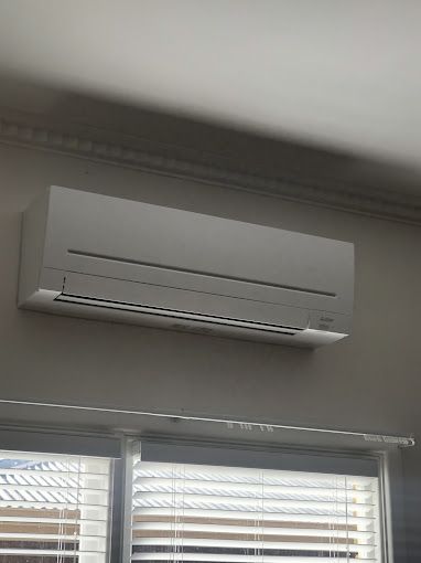 A white air conditioner is mounted on the wall above a window with blinds.