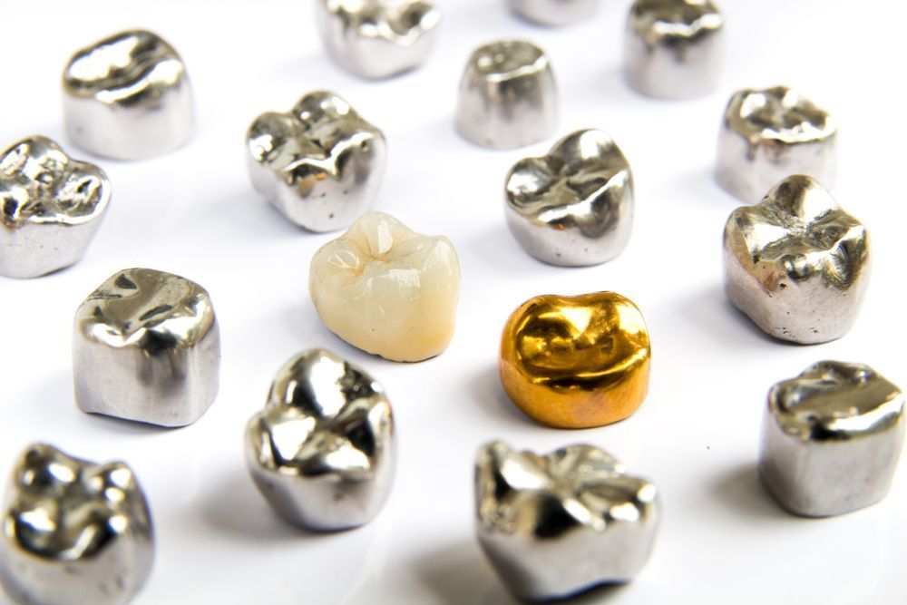 Dental crowns: several silver crowns surround a single, natural-looking tooth, and a gold crown, all on a white surface.