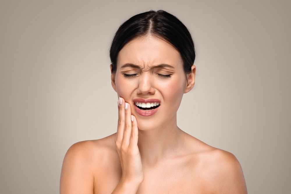 Woman grimaces, holding her cheek in pain, suggesting a dental issue.