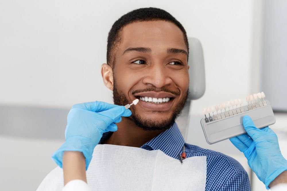 Man in dental chair having his teeth shade matched; dentist holding shade guide, hands in blue gloves.