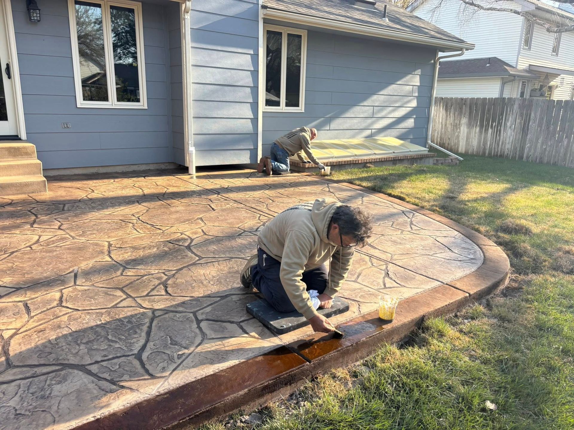 Two men are working on a concrete patio in front of a house.