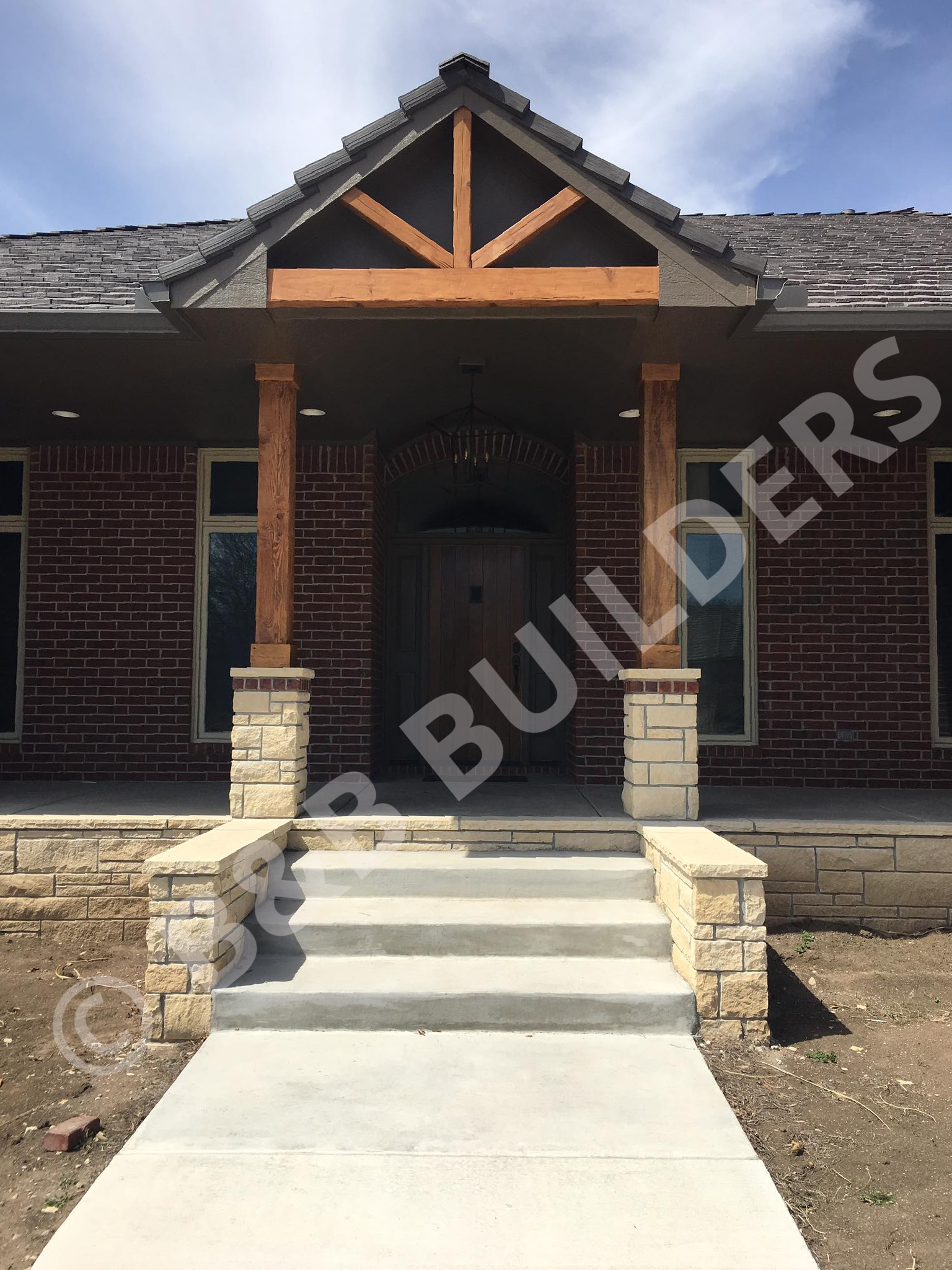An entryway to a house with concrete path and staries leading to a stone porch covered by a timber framed roof