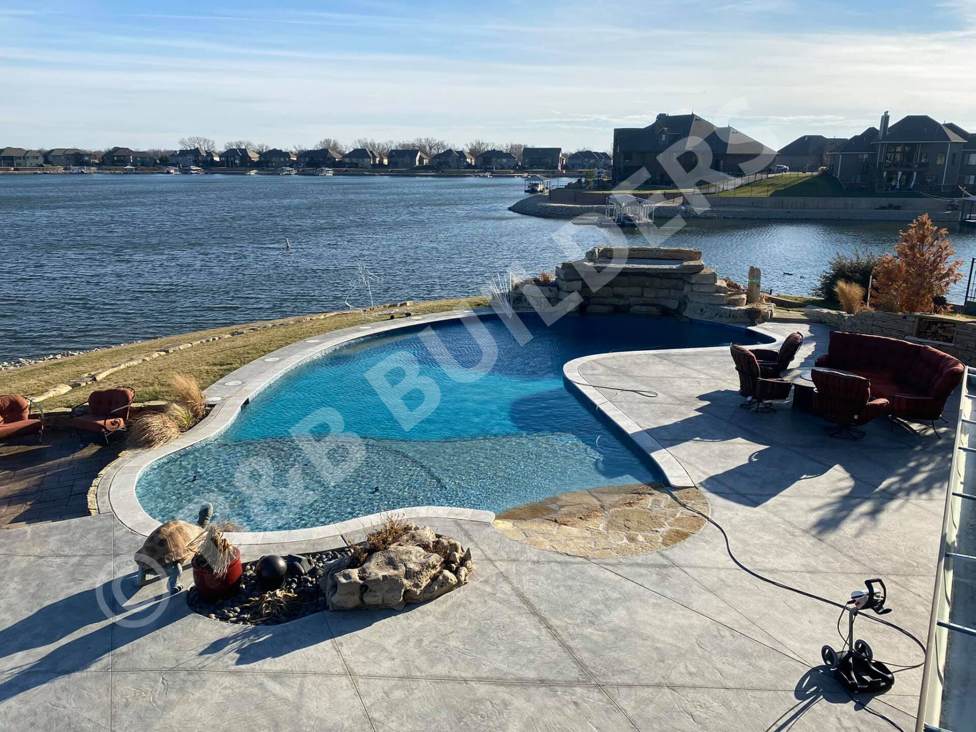 Freeform pool with concrete and stone pool deck, seating area, and poolside stacked stone waterfall, next to a lake
