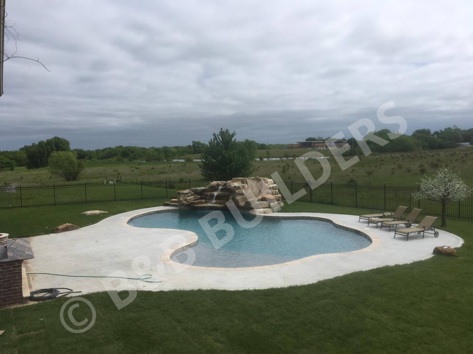 Freeform pool with concrete and stone pool deck, seating area, and poolside stacked stone waterfall with slide
