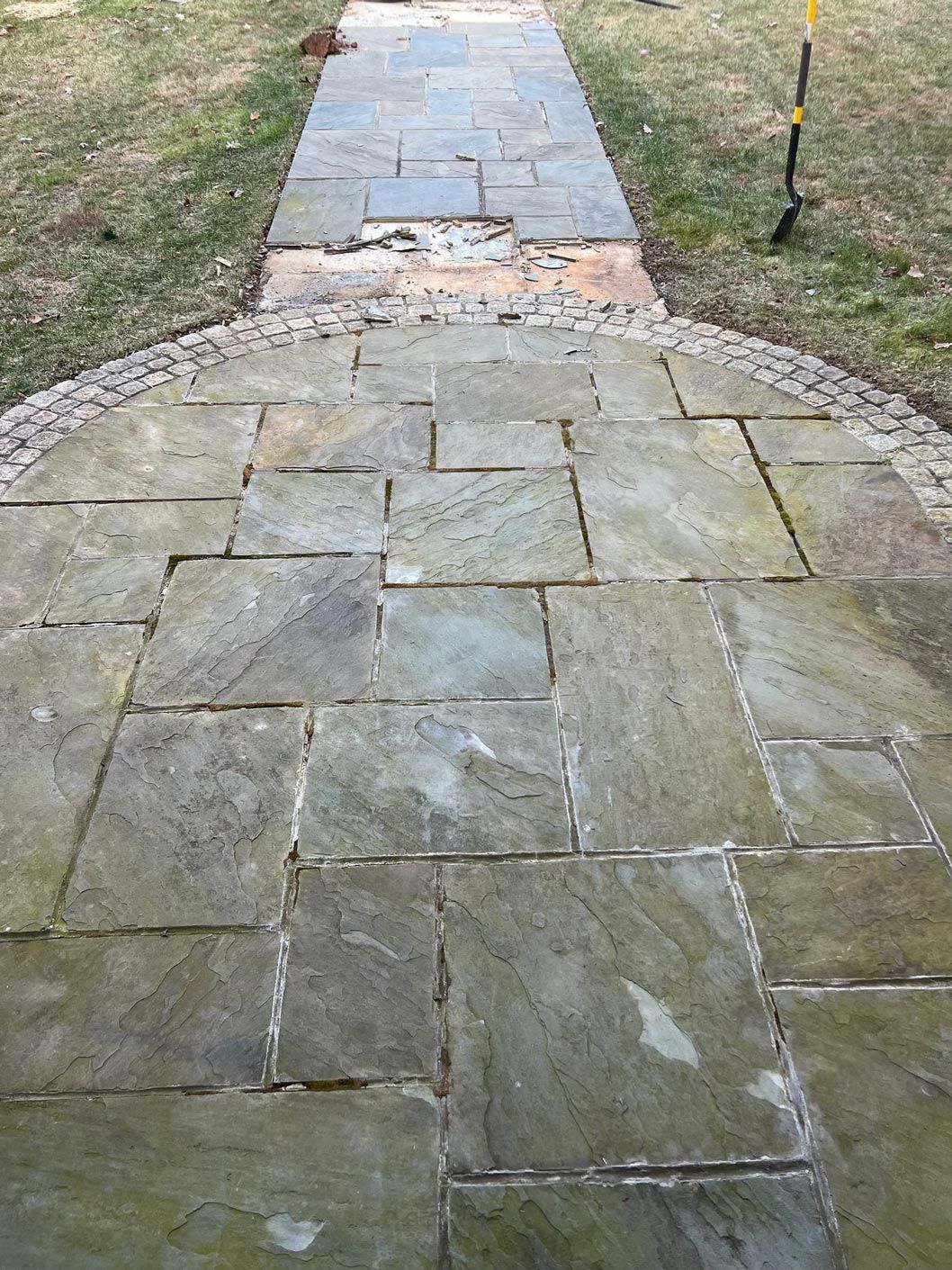 A stone walkway leading to a grassy area with a shovel in the grass.