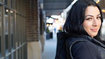 A woman with long black hair (Sarah Delaney of Nirvana Permanent Makeup) is standing on a sidewalk in front of a building.