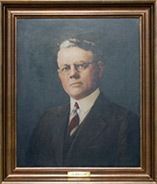 Portrait of a man in a dark suit, glasses, and tie; framed, with a dark blue background.