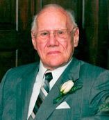Older man in suit, wearing glasses, and a boutonniere, seated indoors.