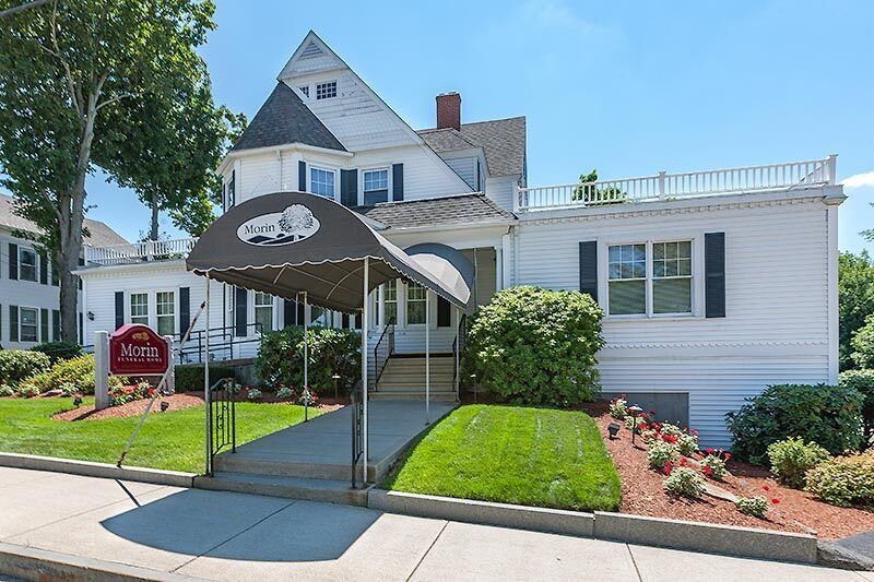 White building with a dark awning, sign, and manicured lawn. Sunny day.