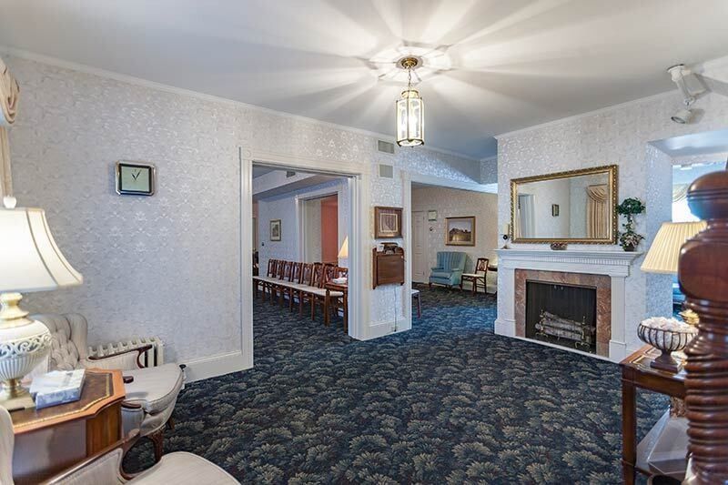 Interior of a funeral home with chairs, fireplace, and ornate ceiling fixture. Dark blue patterned carpet.