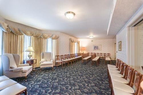 Funeral home viewing room with chairs and curtains. Dark blue carpet, neutral walls, and soft lighting.
