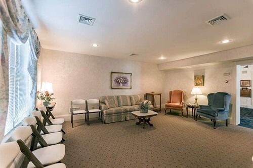 Interior of a funeral home waiting room. Beige carpet, floral sofa, armchairs, chairs, and lamps.