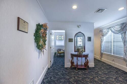 Hallway with floral wreath, dark carpet, arched mirror over table, window with drapes, white walls.