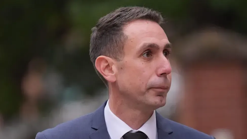 Man in suit, dark hair, looking slightly to the side. Outdoor setting, blurry background.