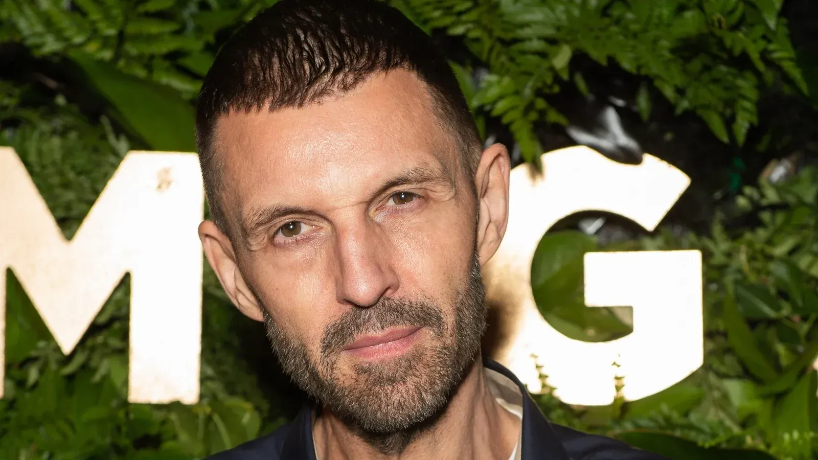 Man with short dark hair, a trimmed beard, and a serious expression at an event with 