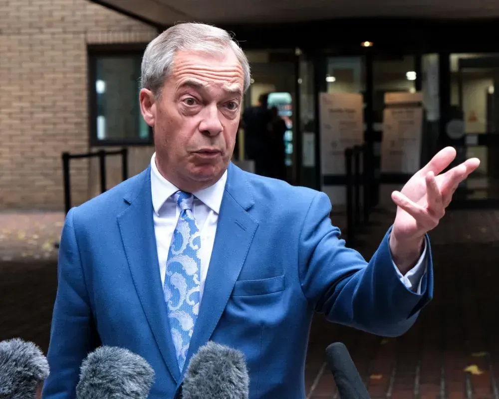 Man in blue suit, gesturing with hands, speaking to microphones outside a building.