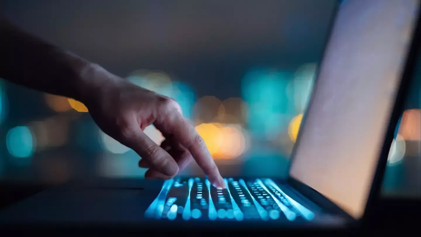 Hand pressing a glowing, backlit laptop key; night cityscape in the background.