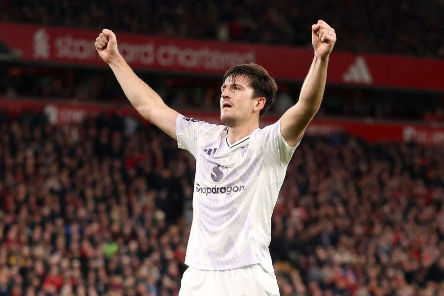 Soccer player with arms raised in celebration, on a field with red stadium seats in the background.