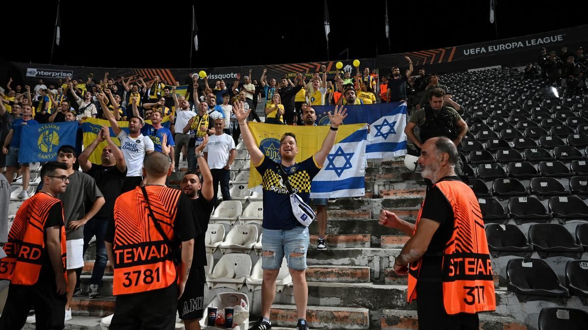 Fans with Israeli flags cheering at a night soccer game; security guards in orange vests are visible.