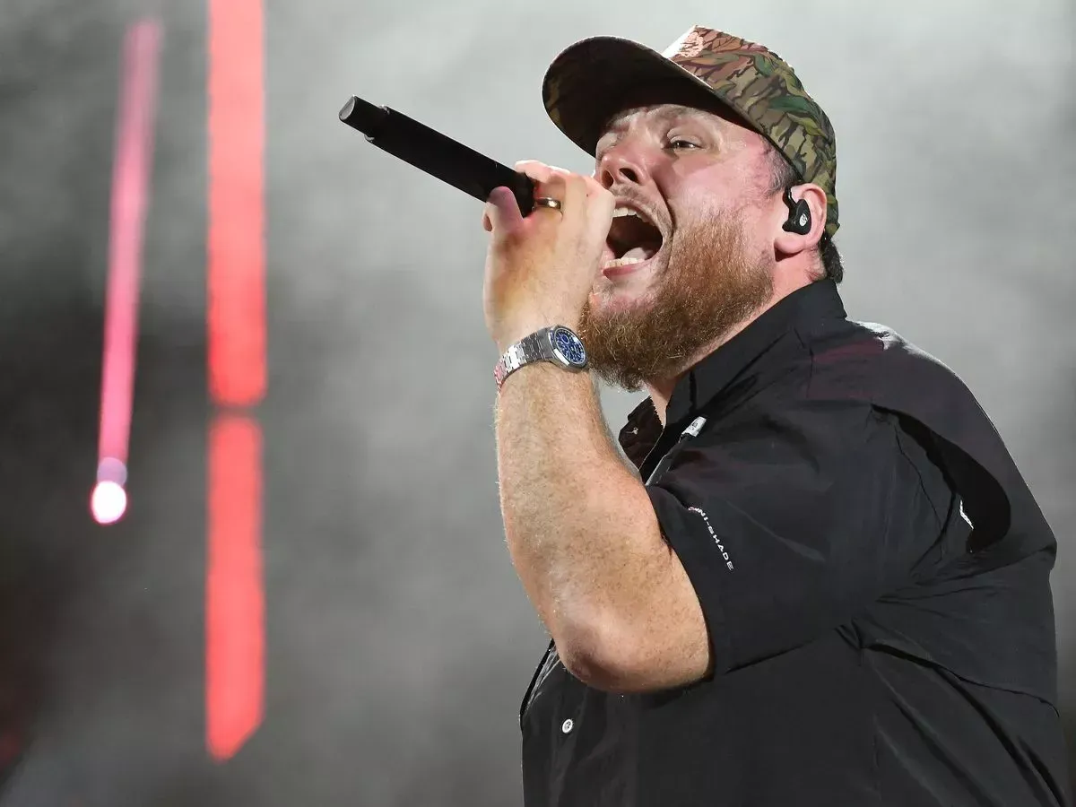 Luke Combs singing into a microphone onstage, wearing a camo cap and watch, with a dark background.