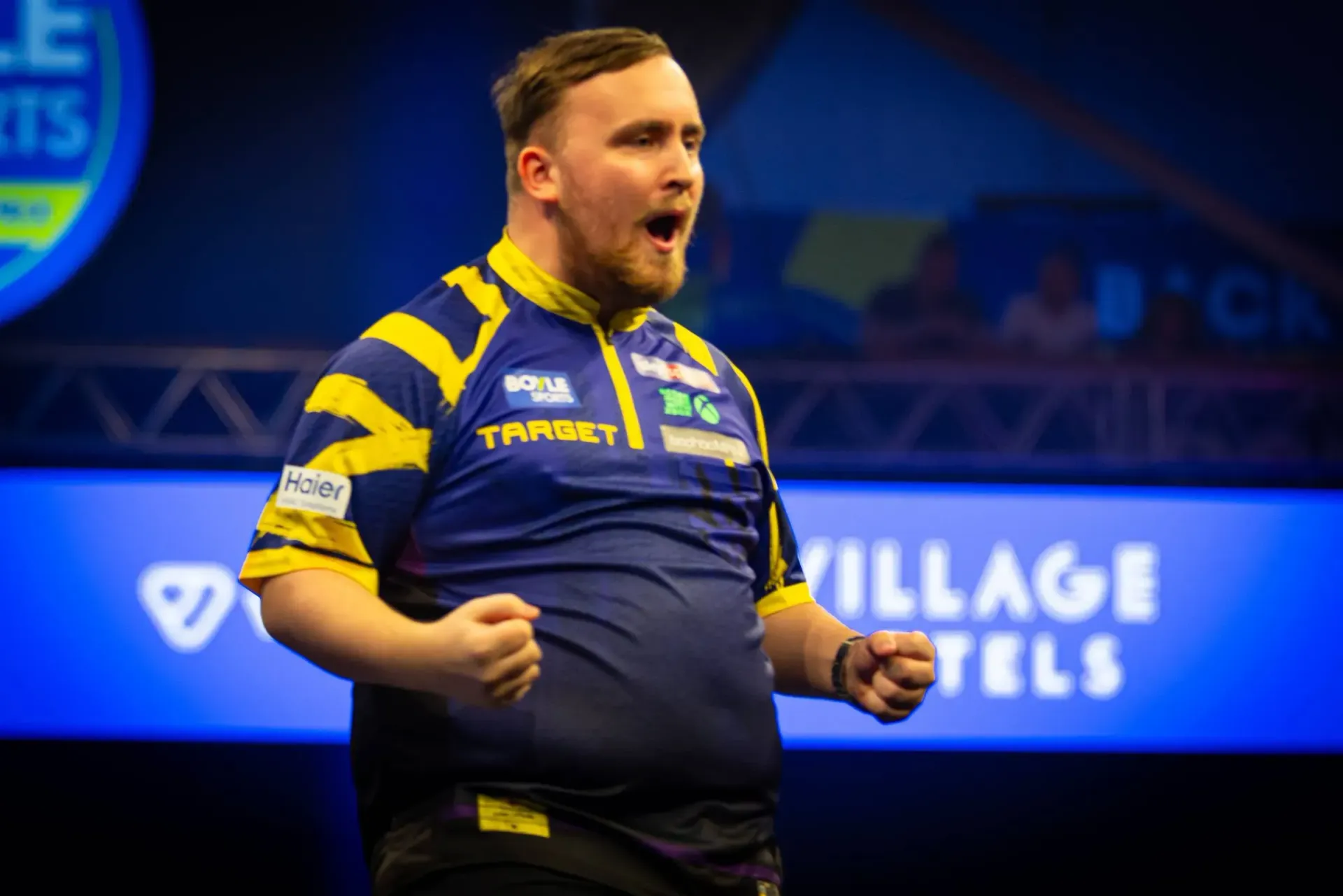 Darts player in blue and yellow jersey celebrates with clenched fists at darts tournament.