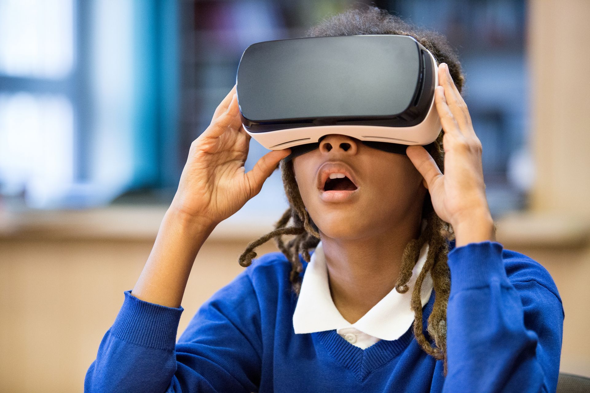 Girl with dreadlocks wearing VR headset, eyes wide open, mouth agape. In a classroom setting.