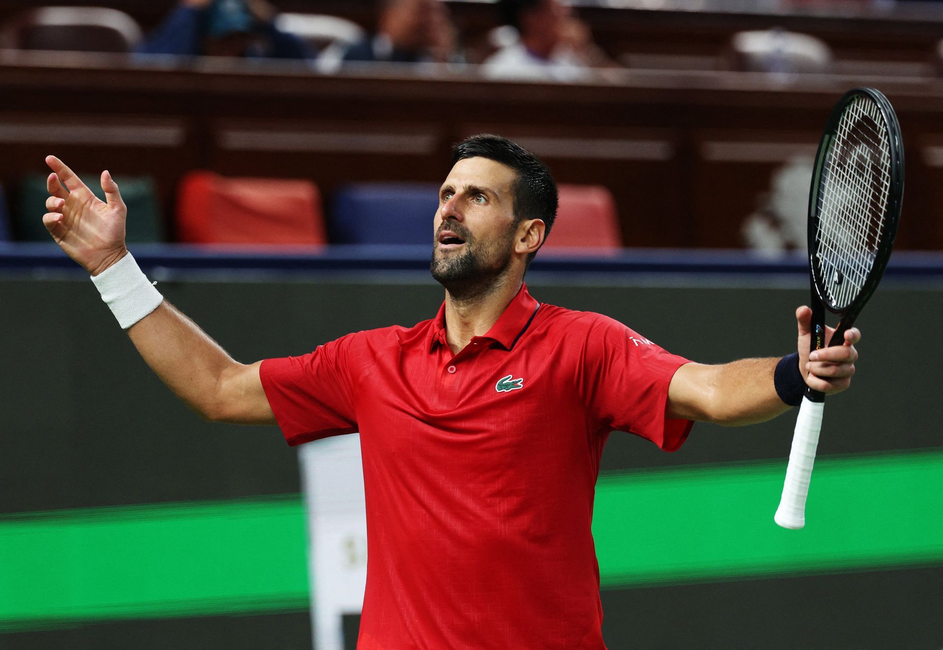 Novak Djokovic celebrates victory, arms raised, red shirt, tennis court setting.