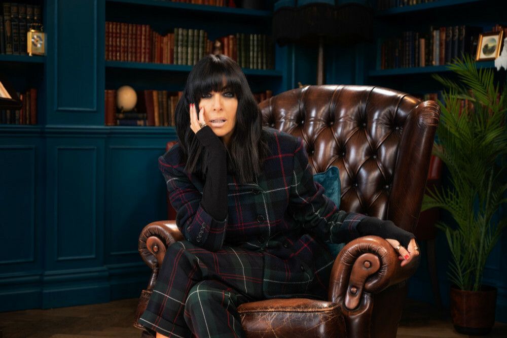 Woman in plaid suit sits in a leather armchair in a library, looking thoughtful.