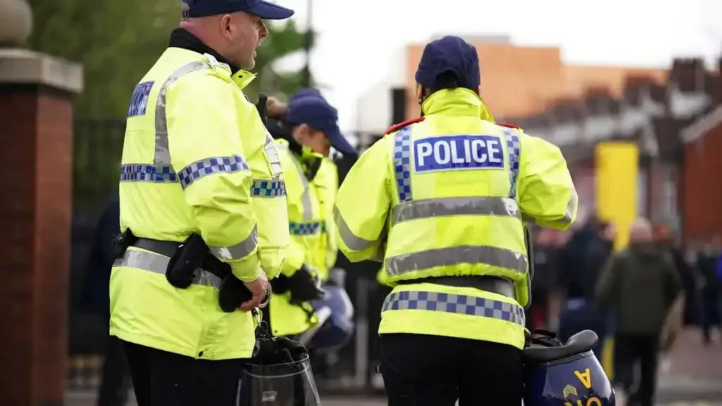 Police officers in high-visibility jackets stand near a stadium entrance; a crowd of people is visible in the background.