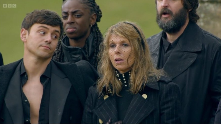 People in black attire at an outdoor gathering. A woman with blonde hair and a man with an open shirt look concerned.