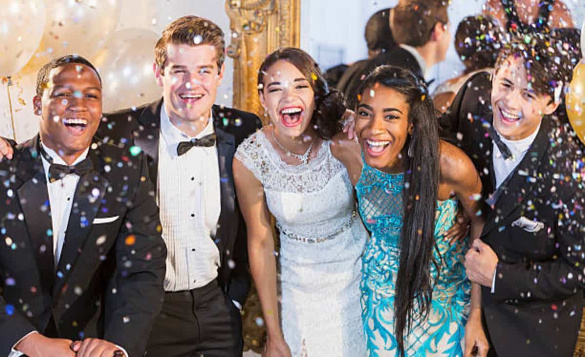 Group of smiling teens at a prom, with confetti falling. They wear formal attire and pose in a well-lit setting.