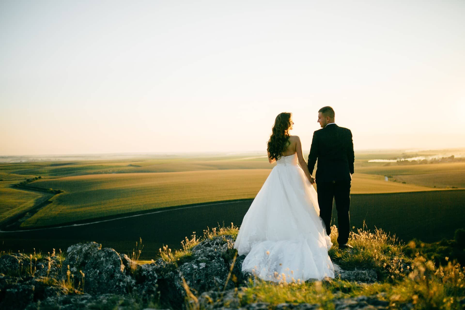 Bride and groom holding hands, facing a scenic sunset over a green landscape from a rocky hilltop.