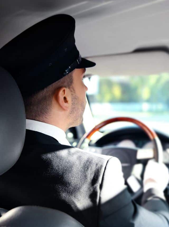 A man wearing a black chauffeur's hat and suit drives a car, seen from behind. He looks forward, holding the steering wheel.