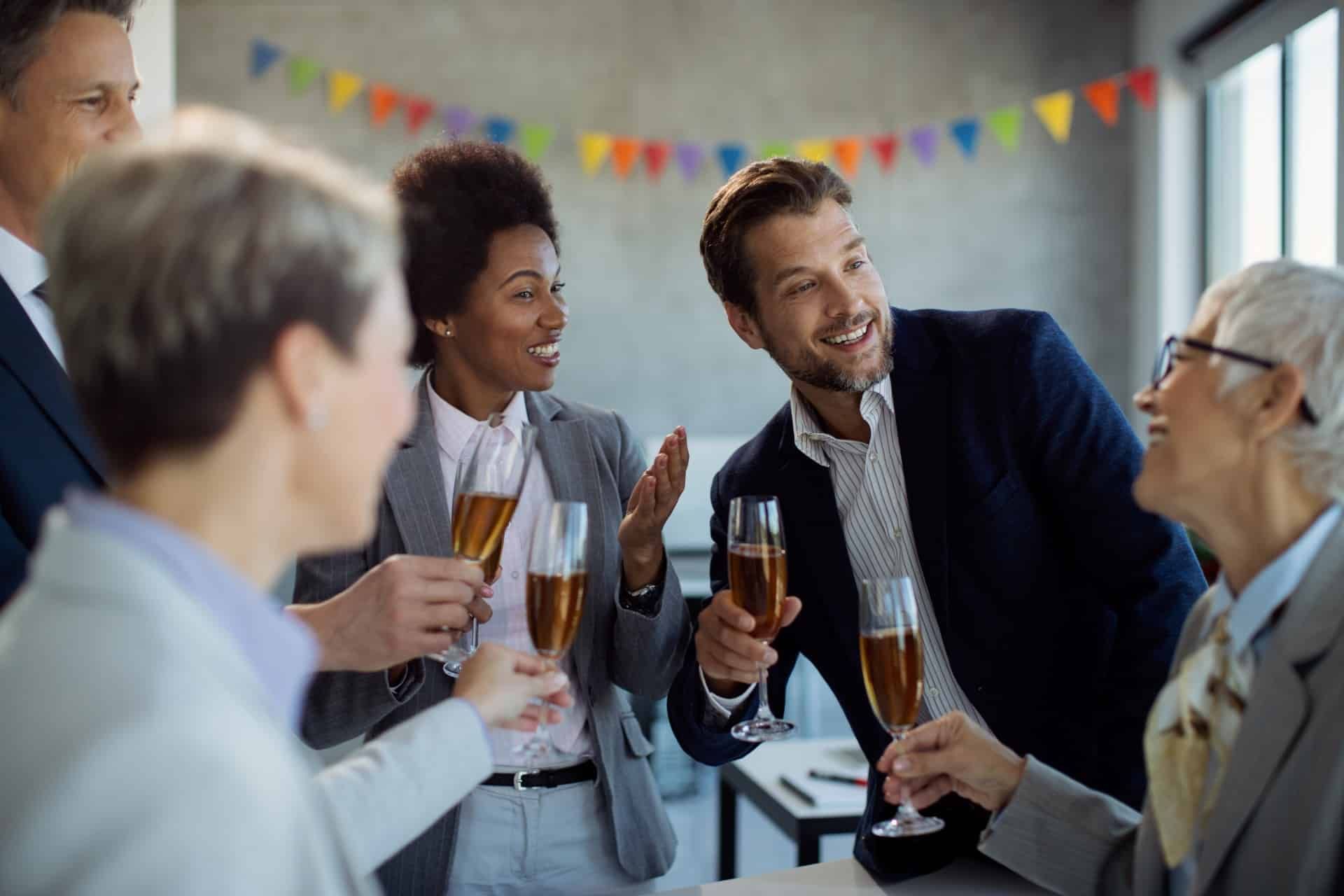 Office colleagues in suits celebrate with champagne, smiling and toasting, with colorful bunting in the background.