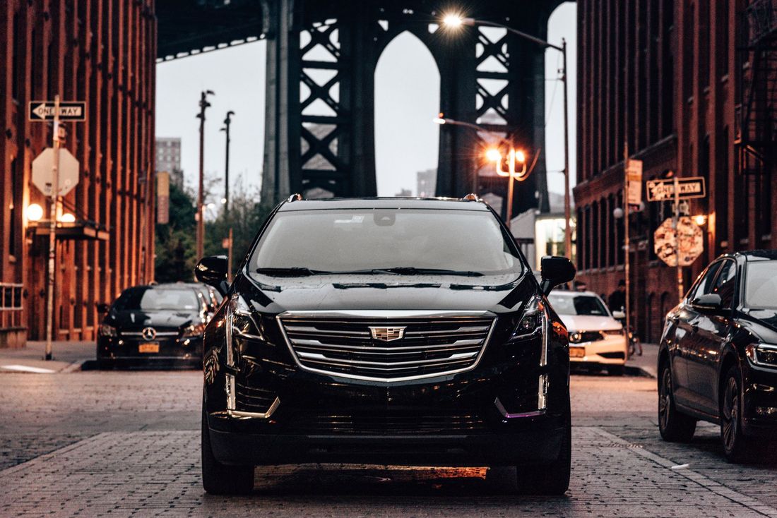 A black Cadillac SUV parked on a cobblestone street in front of the Manhattan Bridge. Other cars and buildings surround it.