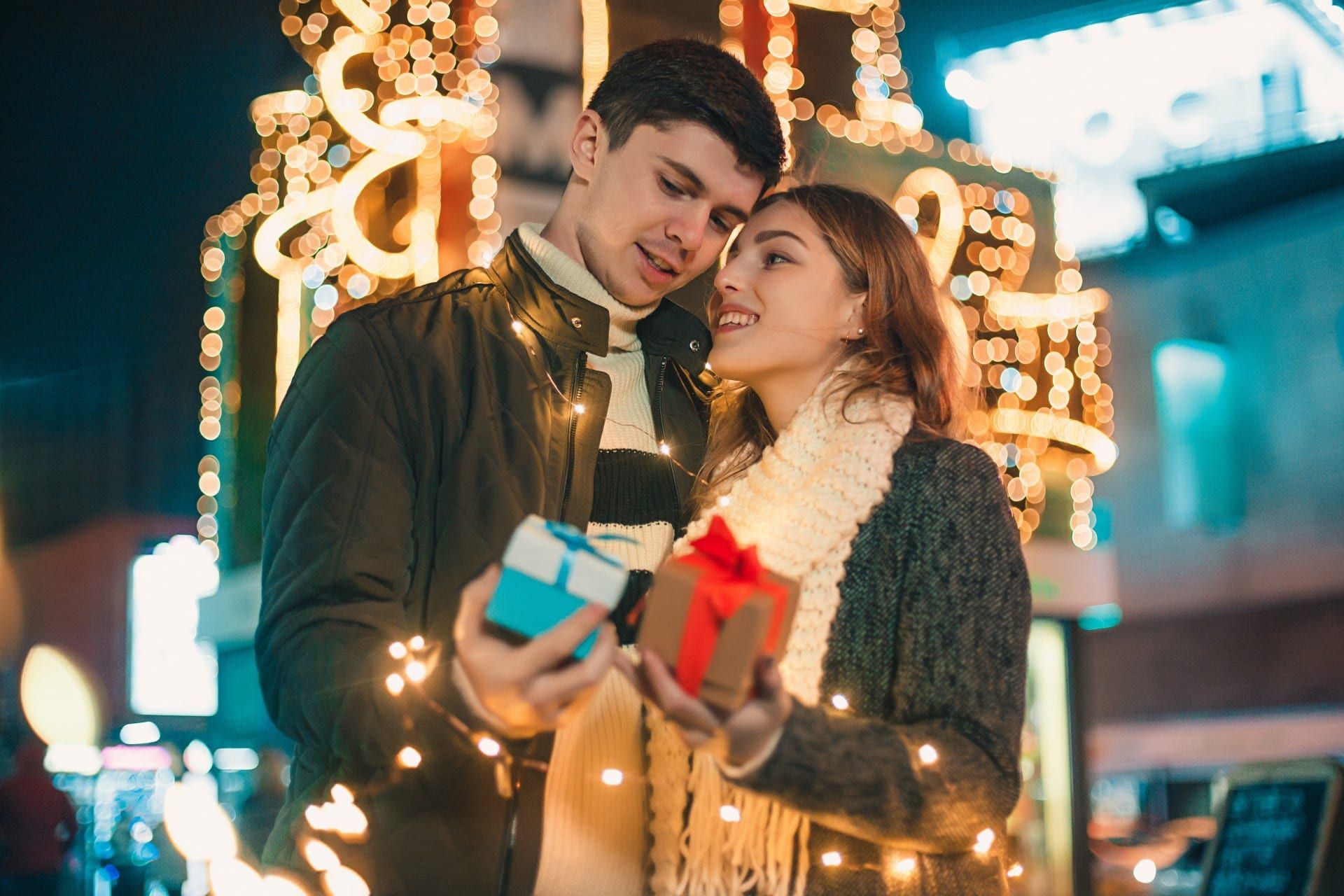Couple exchanging gifts outdoors, illuminated by Christmas lights at night. Man looks at woman, both smiling.