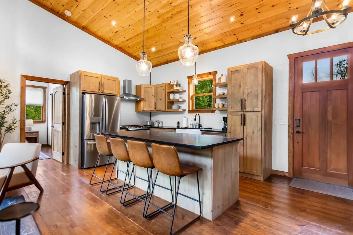 A rustic kitchen with wood-paneled ceilings, a stone island with four leather barstools, and a dark wood front door.