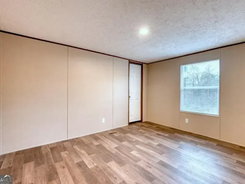 An empty room with light tan walls, wood-look plank flooring, a white interior door, and a single window with blinds.