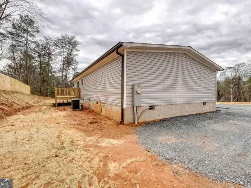 A light gray house with a cinder block foundation, set on a gravel lot next to an unfinished dirt yard and a wooden deck.