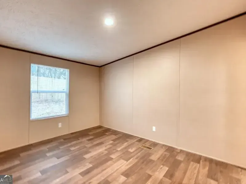 An empty room with tan walls, light wood-look flooring, a ceiling light fixture, and a window with white blinds.