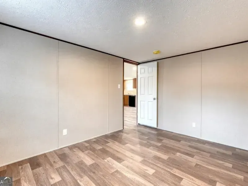 Empty room with light-colored walls, wood-look flooring, and an open white door leading to a kitchen.