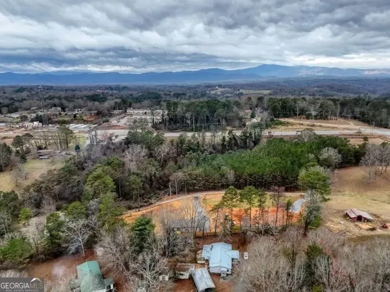 Aerial view of a rural landscape with a farmhouse, outbuildings, surrounding woods, and distant mountains under a cloudy sky.