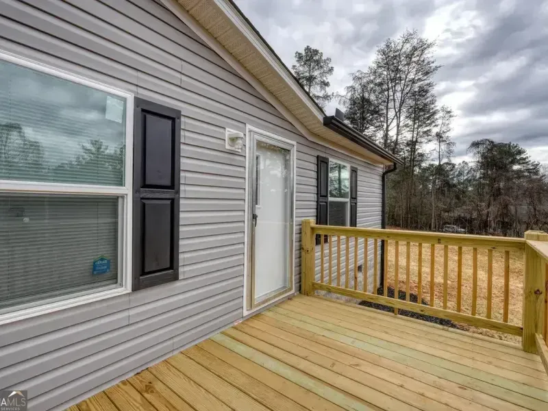 A wooden deck with a railing attached to the side of a light gray vinyl-sided house with a white door and dark shutters.