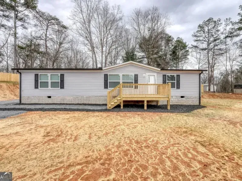 A light gray, single-story manufactured home with a wooden porch, set on a plot of land with bare ground and trees.