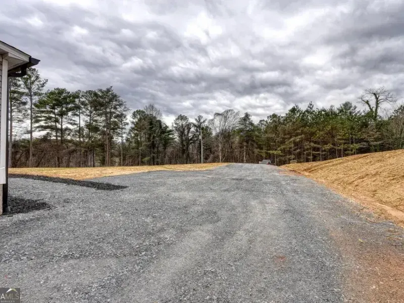 A wide gravel driveway leads toward a tree line under a cloudy sky, with a portion of a building visible on the left.