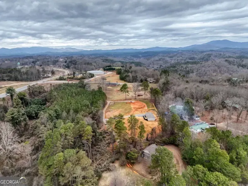 An aerial view shows a rural hillside with patches of red earth, scattered trees, a house under construction, and mountains.